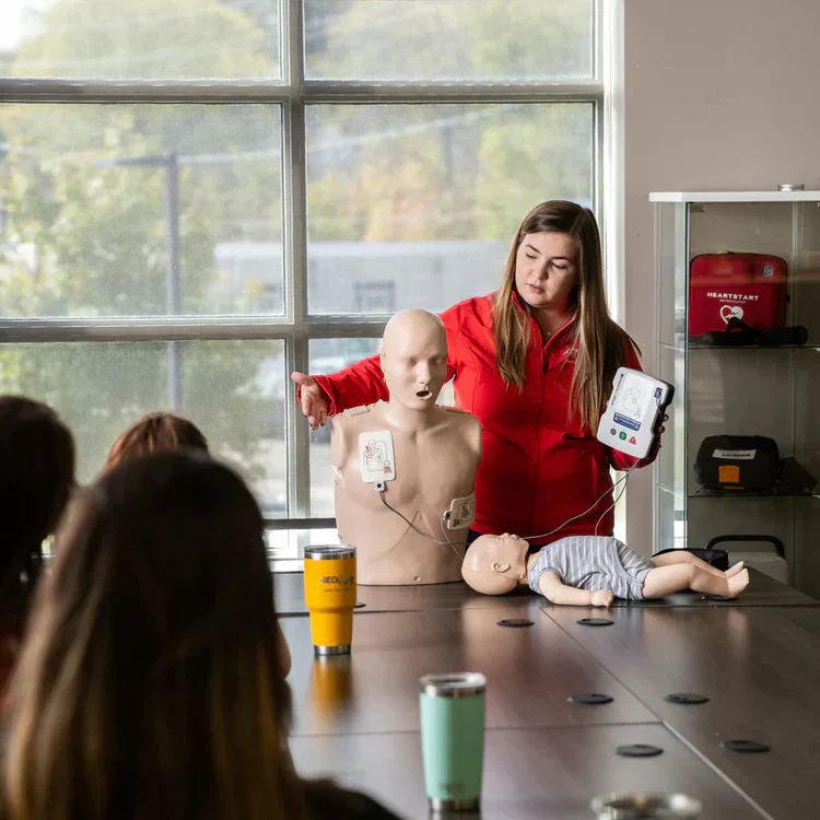A woman teaches CPR using a mannequin and AED device to a group seated around a table.